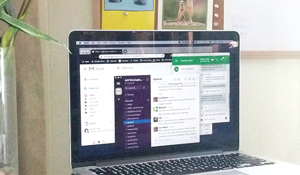 A MacBook Pro with Gmail, Hangouts, and Slack all open, on a glass desk in front of a bulletin board